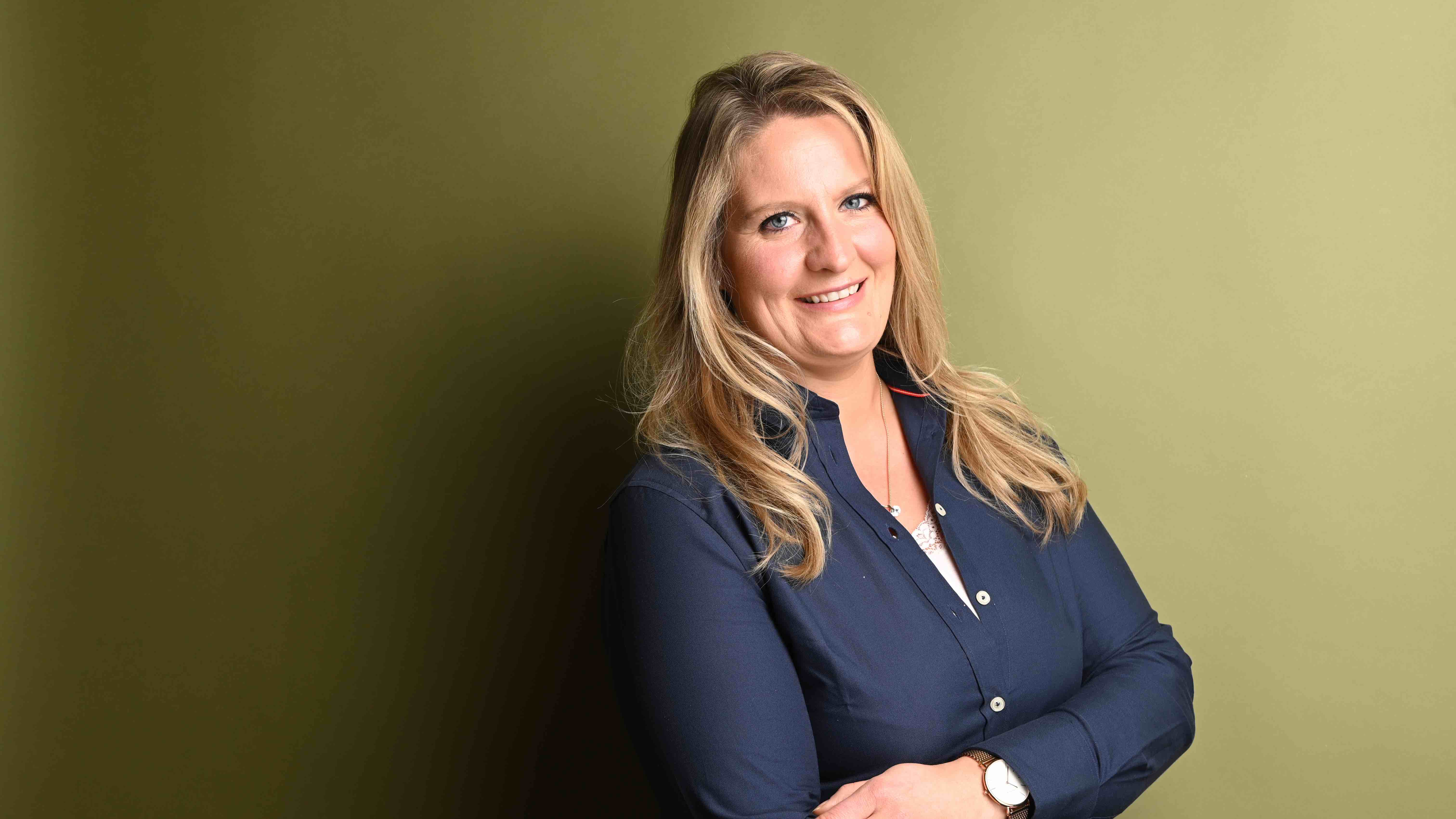 Lächelnde Frau mit Brille steht draußen in der Nähe blühender Büsche.