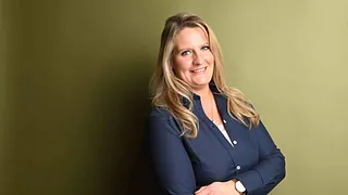 Lächelnde Frau mit Brille steht draußen in der Nähe blühender Büsche.