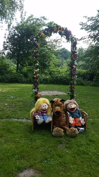 Drei Puppenfiguren sitzen unter einem floralen Bogen im Freien.