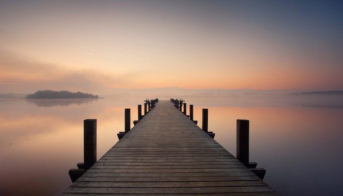 Holzsteg erstreckt sich über ruhiges Wasser in Richtung nebligen Horizont.